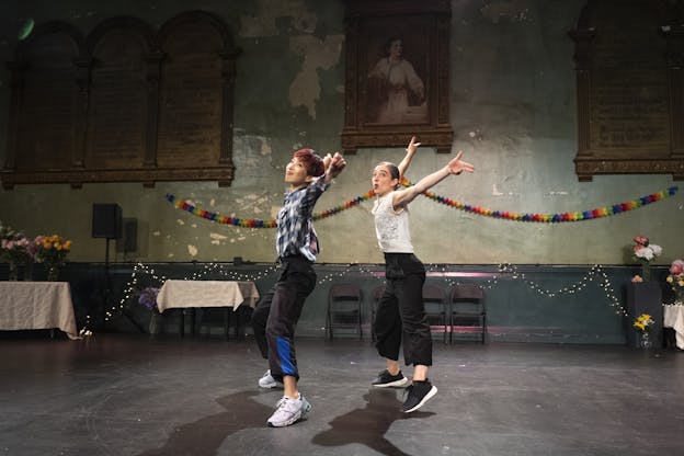 Two performers dance in the middle of a space decorated for a party. The two dancers are centered in the frame, both facing the left and standing in jumping jack positions with their feet wide apart and arms raised, their heads are turned to look at the camera. Hsiao-Jou Tang dances on the left half of the frame with a smile on her face, gazing above the camera and making snaps with her hands. She is wearing a blue and white flannel button up, black sweatpants with blue stripes above the cuffs, and white sneakers. Monica Bill Barnes dances on the right of the frame, also gazing upwards. She is wearing a white sleeveless blouse with a lace front, black high-waisted pants, and black sneakers.