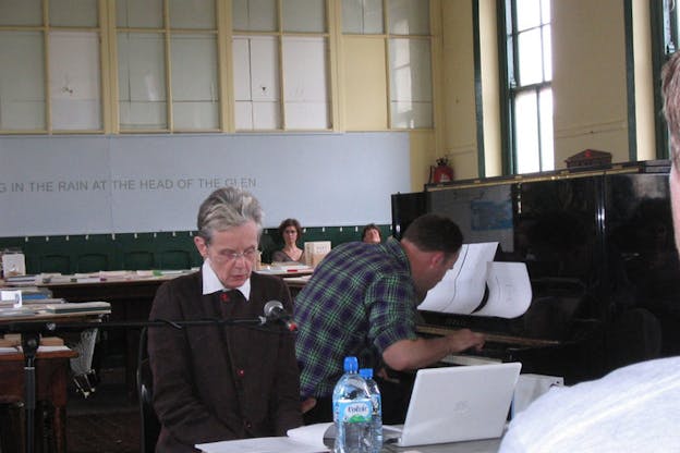 A performance still featuring one individual speaking into a microphone secured on a horizontal arm and another individual playing the piano. Two other people sit and watch them from the back. They appear to be in a classroom setting with yellow trimming around windows on the back and side walls and blue cut off text on the back wall reading 