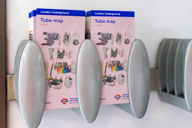 A round metal pamphlet holder is bracketed to a white wall and holds two stacks of light pink pamphlets side by side. A small blue banner at the top of the pamphlets reads 