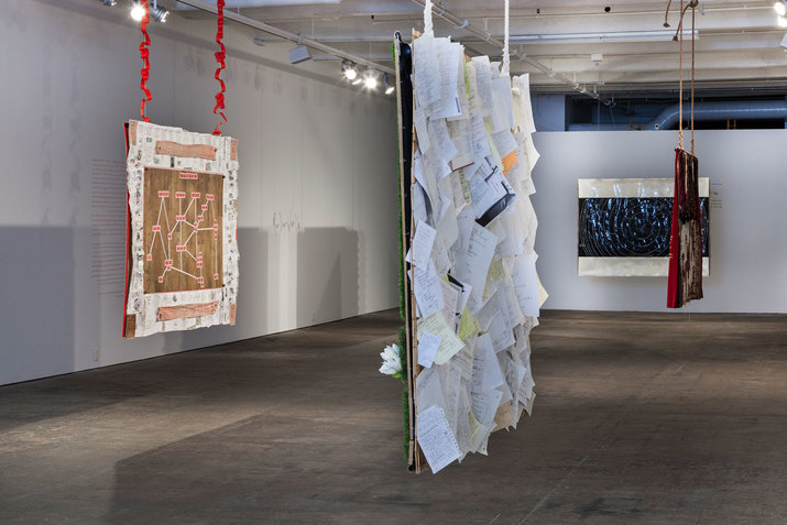 Gallery installation of four hanging quilts, one arranged with loosely bound pages, one with a dark blue spiral diagram, one with a straight-line diagram, and one with red cloth and mixed-fabric. 