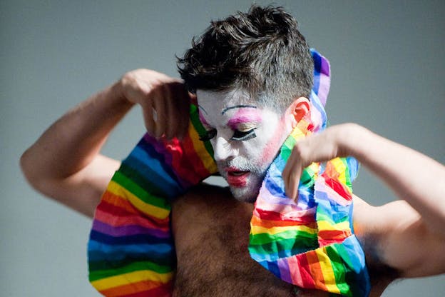 Close-up of a bare-chested performer wearing white face paint, pink painted eyes, fake eyelashes, red lipstick, and red blush drapes a rainbow-colored piece of fabric behind their neck.