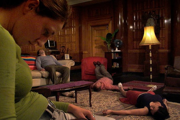 Photograph of four people in a room in various positions. One of them is seated down on a couch staring at the ceiling, while the another lays down with feet propped on a red armchair, with another person laying right next to him. In the front, closest to the viewer another person is seated staring down.