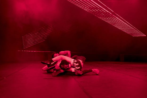 Three figures lay on the ground tangled on top of each other, with two facing the front and the third an upside down direction. The room they are in lighted red.