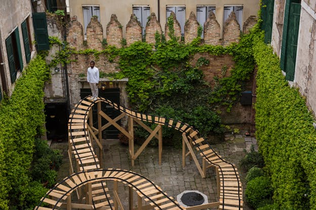 Eagle's view image of a figure clad in white standing on top of a  wooden structure similar to a fun park's train rails. The structure is elevated by wooden sticks in the middle of a stone backyard of buildings.The buildings are covered by green shrubs creeping on the bottom of their walls. 