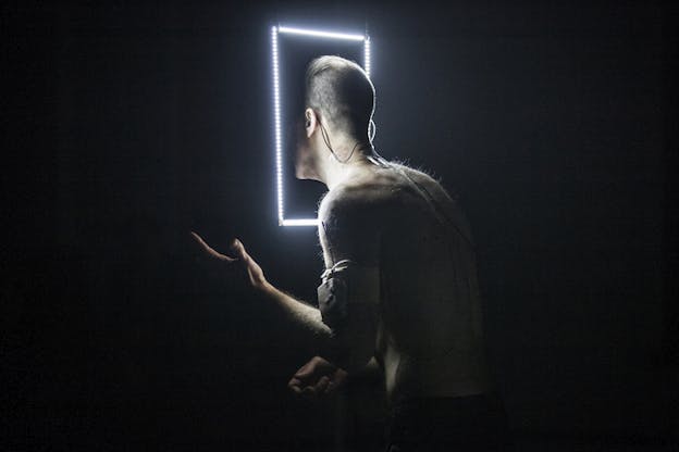 A shirtless figure standing sideways in black surroundings emerges half of their face in the dark inside a hanging white lighted frame. 