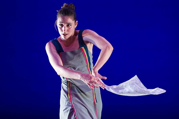 A performer in a gray dress with two rainbow stripes on each side holds a handkerchief in front while moving.