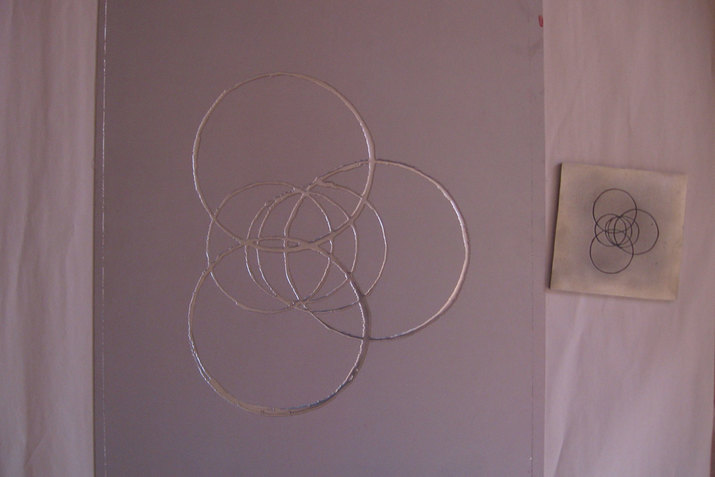 Silver intersecting circles in an intricate venn diagram shape on a mauve-colored board photographed on a bed beside a stamp of the same circle design.