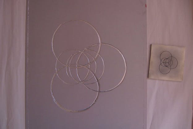 Silver intersecting circles in an intricate venn diagram shape on a mauve-colored board photographed on a bed beside a stamp of the same circle design.