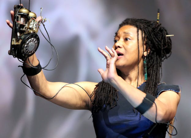 A photograph of Pamela Z performing with her arms upraised up, holding a rotary from a phone, with the wires from it circling her arms. The background of the image is lavender and blurred. Pamela Z wears a blue top and green dangling earrings.