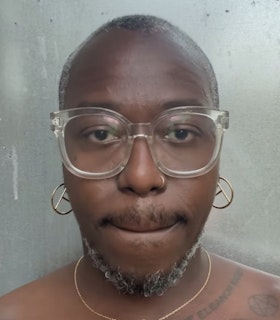 A portrait of Brontez Purnell against a light grey wall, wearing clear frame glasses, a silver nose ring, and silver hoop earrings. His hair is short and he has a short black and grey beard. A few tattoos are visible on his chest. 
