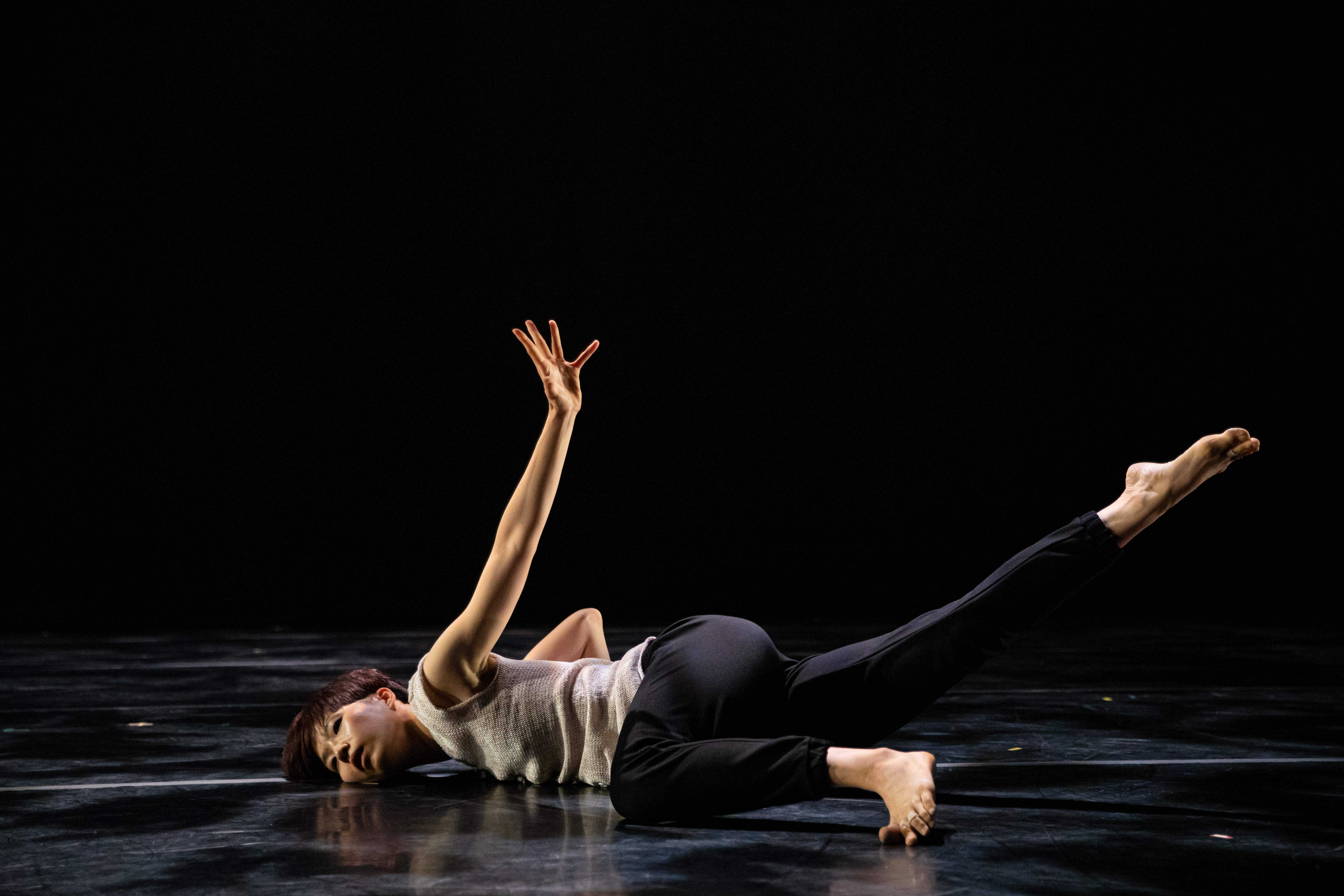 Hsiao-Jou Tang is centered alone as she dances on a dark stage illuminated softly from above. She lays on her stomach with her head pointed to the left and her feet on the right half of the frame. Her head is twisted for face the camera, she looks out, blankly. Her left arm is extended straight back, hand reaching abover her with outstretched fingers. Her left leg is bent touching the floor, and her right leg is kicked straight out behind her. She is barefoot and dressed in a light grey, knitted sleeveless top and black pants with elastic cuffs.