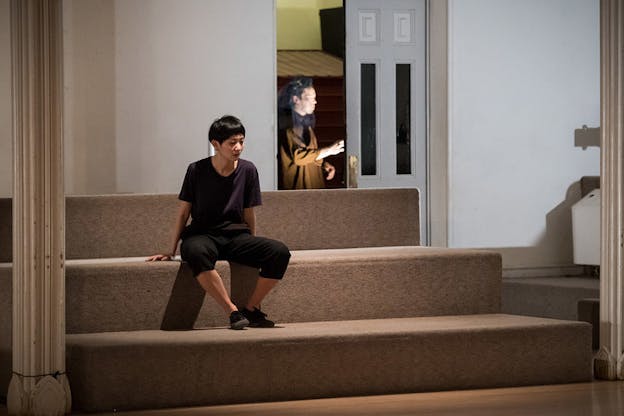 A performer clad in black sits in the middle of three stairs looking in the distance. Behind them in the distance a figure dressed in black with a transparent black hued veil pushes on a door. 