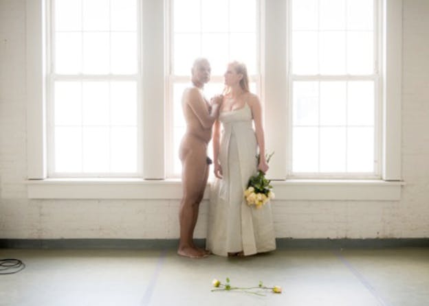 A person in a white gown holds a bouquet of yellow roses and turns their face to a fully nude person standing sideways with their arms on the clothed persons' shoulder.  They stand against a white painted exposed brick room with tall drawn windows and soft light. 