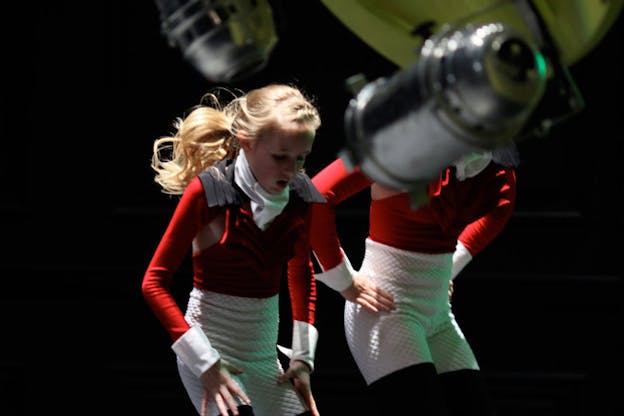 Two performers stand with their hands near their waist and their hips bent. The face of one performer is visible and they have an expression of determination. Mechanical objects attached to a lime green object obscures the view of the other performer's face.  