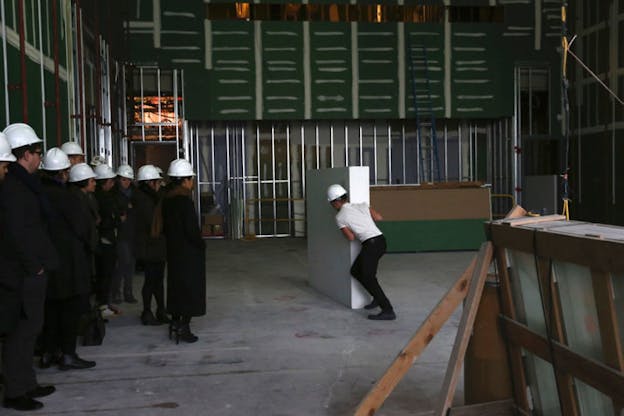 A person dressed in black pants, white shirt and white construction hat pushes a white rectangle box as a crowd dressed in black with white construction hat watches from the left.  