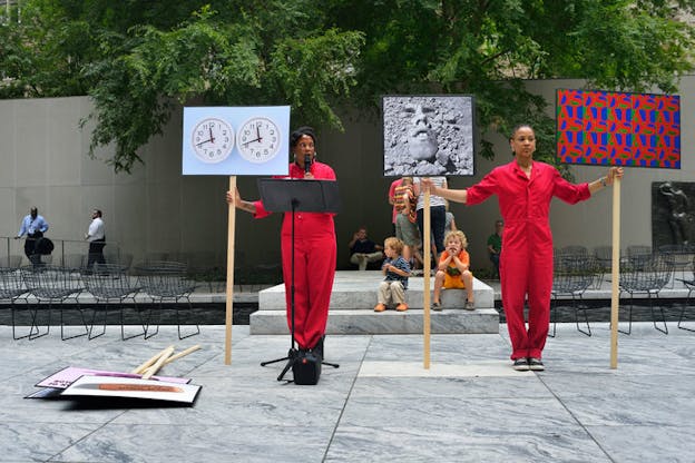 Two performers in bright red jumpsuits hold three signs: one with abstract lettering repeating 