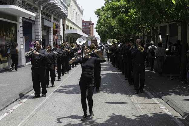 People dressed in black and playing instruments form three parallel lines on a street while being led by a person standing in front of the middle line holding a cane.