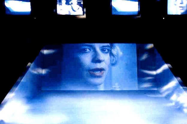 A projection of a woman's face on a smooth concrete surface, with two declining walls on the side. Everything is covered in sea blue light with small reflections of white.