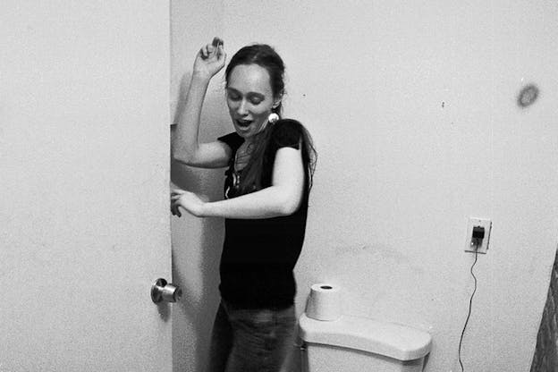 Black and white photograph of person dressed in jeans, a dark shirt and circular earrings in a bathroom leaning against the wall near the door.