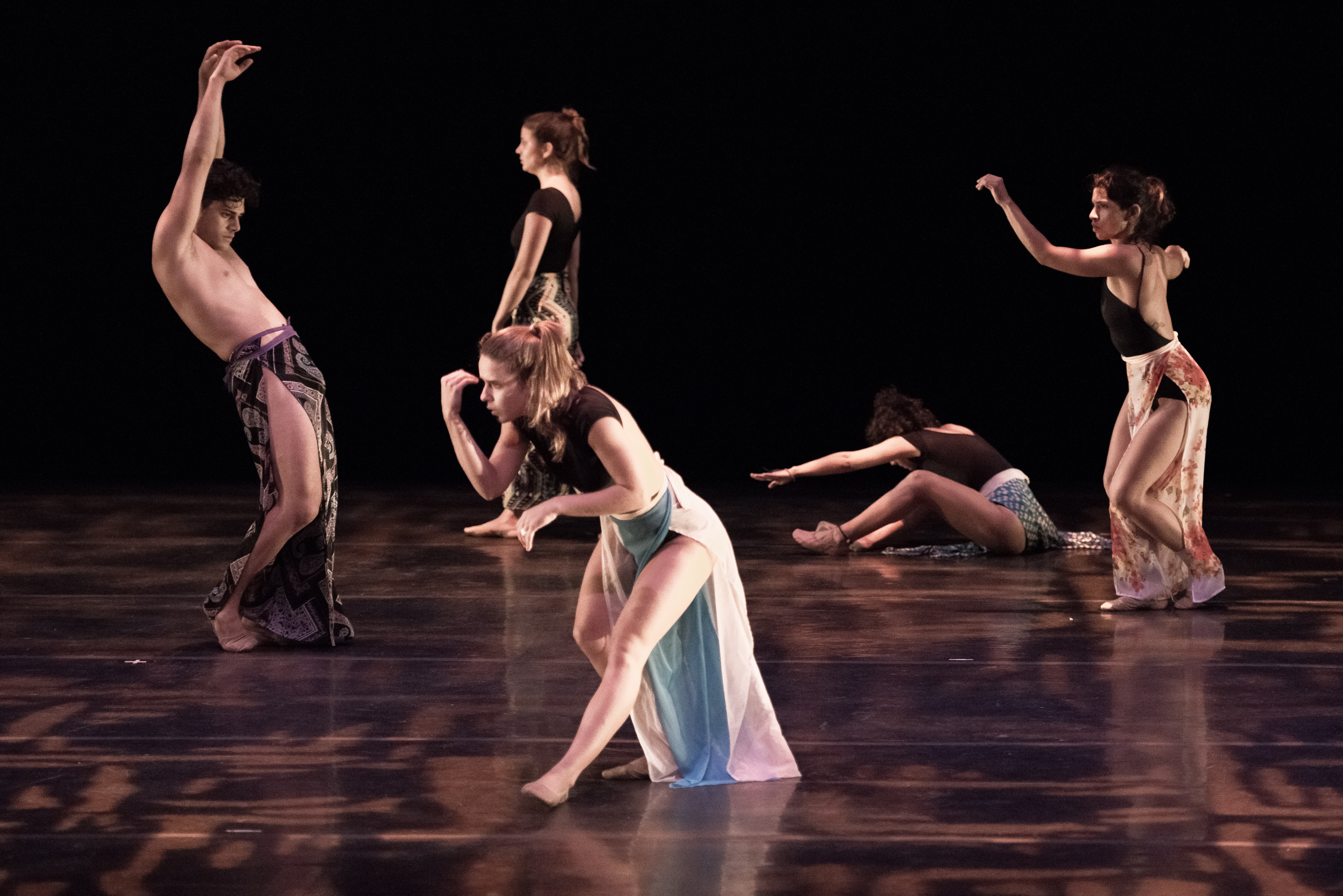 Five dancers perform on a stage lit by soft orange and purple light, dressed in long patterned colorful skirts and leotard tops. Four are posed in various positions in a small arc towards the back of the stage, and the fifth is centered, standing closer to the front of the stage, and poses facing towards the left. 