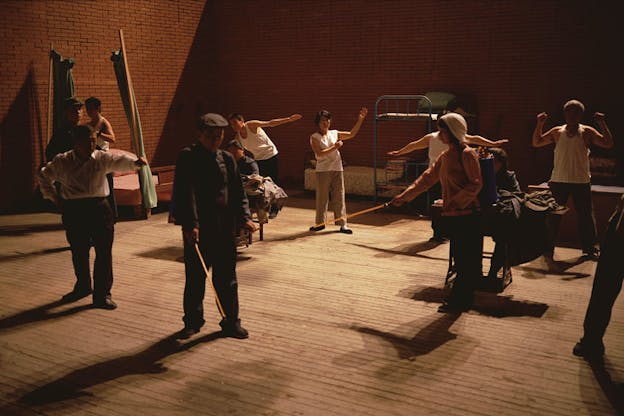A performance still of several performers in a room with red brick walls and various cots against the walls. The performers wear street cloths and stand in various positions suggesting interrupted action. Two performers weild wooden rods while others extend their arms out in front of them. All of the performers look resolutely ahead. 