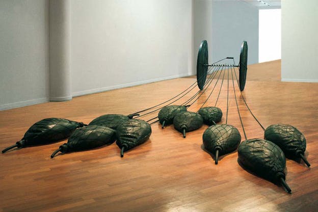 Two thin tires are joined by a tube attached with bags of sand and water secured in nets. The installation sits on a wooden floor against an off-white background.  