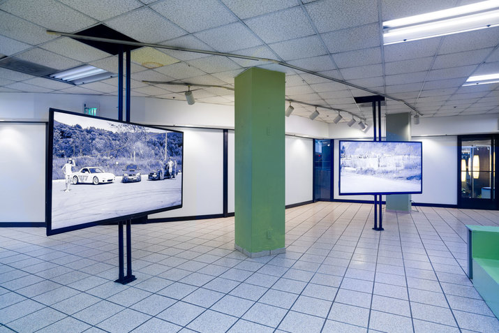 Two suspended screens displaying blue hued images of a fence growing amidst vegetation and people standing beside parked cars in a rural lot are separated by a mint-green column. 