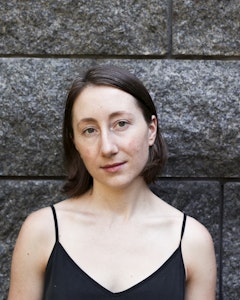 Portrait of Rebecca Hadley dressed in a black tank top, with short brown hair parted in the middle and green eyes standing in front of a gray wall.