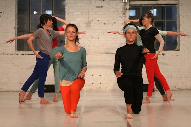 Two persons dressed in dancewear lunge forwards with one arm outstretched while behind them, two rows of dancers face each other with their arms and legs alternately raised and pointed.