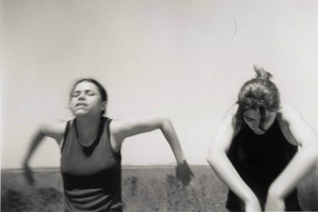 A black and white performance still of two performers wearing dark colored tank tops. The performer on the left closes their eyes and loosk forward with their arms bent at a ninety degree angle behind them. The performer on the right looks downwards with their arms bent at a ninety degree angle in front of them. In the background, there is a clear sky and a grassy field. 