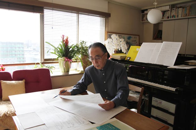 A person in glasses and a gray button up seated with their back to a piano on a wooden table, looks down on music sheets scattered around the table.