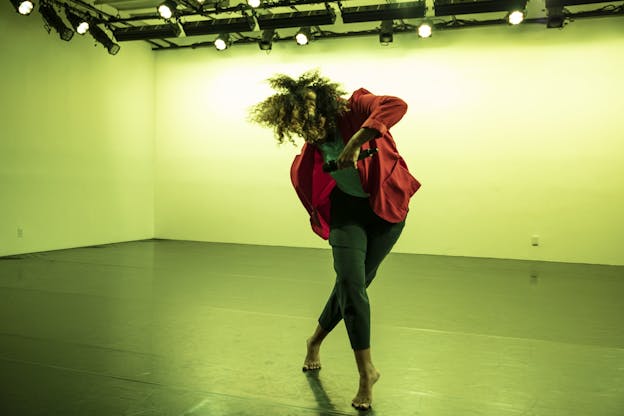 A performer stands alone in a space lit with lime green light. They wear a red jacket and grey pants and are barefoot. Their head is angeled downwards and away from the viewer. Their legs are crossed and their left arm is raised and bent. 