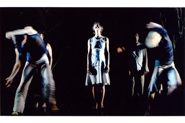 Three performers wearing light blue clothes stand in a black space and face the camera. In front of them, two other performers wearing darker blue clothes are blurred. The performer on the left leans forward and the performer on the right leans backwards. 