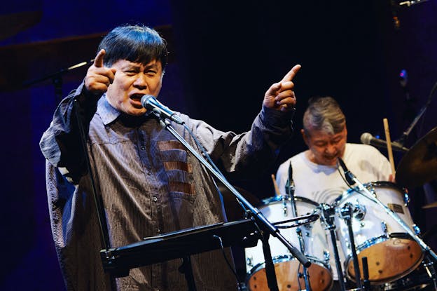Koichi Makigami performs on a lit stage with a drummer behind him who is slightly out of focus. Makigami stands on the left side of the frame facing the camera. He has a theremin, a tablet, and a microphone in front of him. He looks towards the camera as he vocalizes into the microphone, his hands are raised above his shoulders as he points outward with his index fingers. He is wearing a dark, oversized striped button up shirt. 