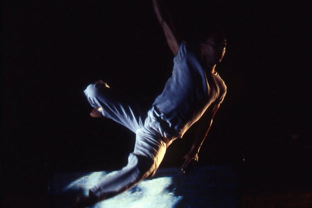 A performer wearing a blue t-shirt and blue jeans leaps within a black background. Their left arm extends fully out behind them and their right arm hangs low at their right side. Their legs are both bent at the knee. A blurred light blue project is beneath them.