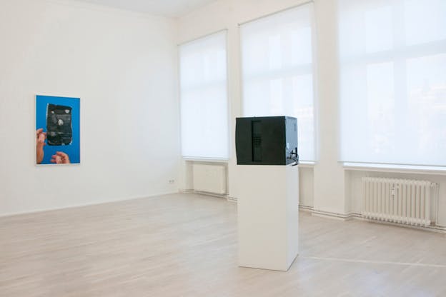A gallery room with a white rectangle supporting an electronic box with its back to the viewer. In front of it an image on the wall with two hands holding a black item on a blue background.