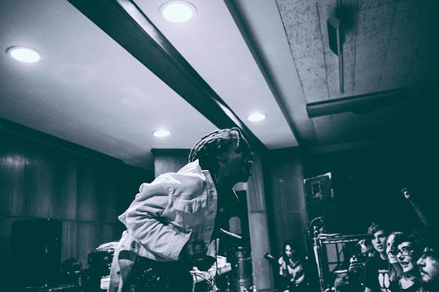 Grayscale photograph with purple undertones depicts a performer in a white jacket singing while an audience underneath watches in amazement.