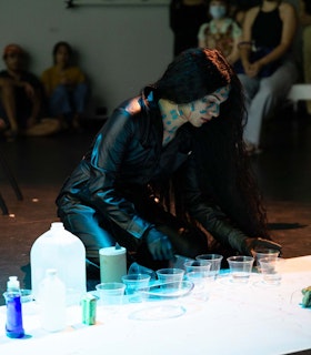 Angel Lartigue kneels on the floor looking down at several plastic cups of clear liquid. She has blue paint speckled on her face and neck. She wears a black leather one-piece outfit with black surgical gloves and hoop earrings.
