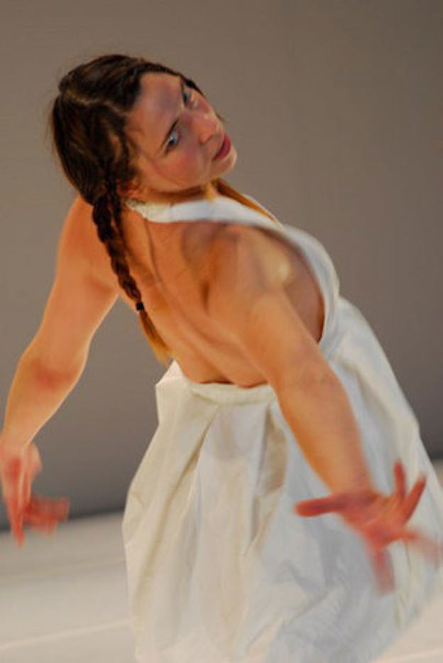A performer wearing a white dress leans backwards, turning their head to the right and extending their arms downwards and behind them. 