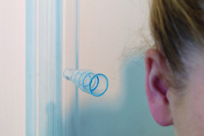 Close up shot of a person's ear on the right side with a tubelike transparent piece. The piece whose rim becomes bigger closer to the ear hangs in front of a white wall with a streak of blue paint.