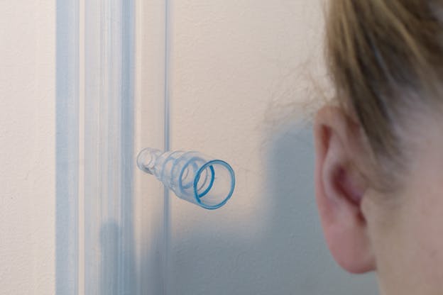 Close up shot of a person's ear on the right side with a tubelike transparent piece. The piece whose rim becomes bigger closer to the ear hangs in front of a white wall with a streak of blue paint.