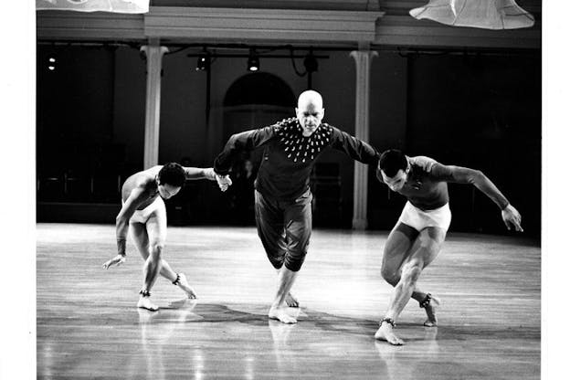 Black and white image of three barefooted performers hold hands while their left leg, situated in front of their right, bends. The two performers on the right and left have their bodies and head facing towards the ground.