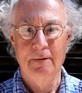 A close up portrait of David Behrman looking slightly below eye level. He has white curly hair and wears a pair of glassed and a blue flannel shirt.