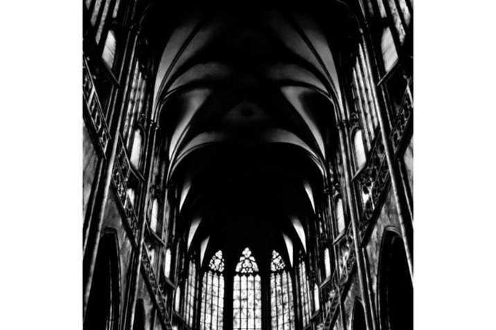 Slightly blurred black and white photograph of a vaulted cathedral ceiling with tall arches, stained glass, and ornate walls.