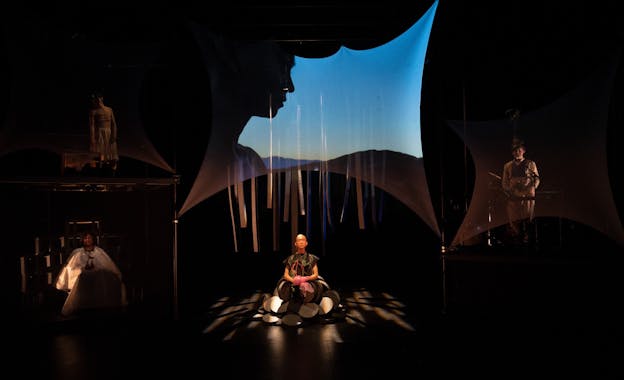 eddy kwon sits illuminated in the center of a dark stage half-buried beneath a heap of circular objects. Three other performers are scattered across the stage around her. Behind and above her head, a face silhouetted against a sky at dusk is projected onto a stretched screen. Behind the projection, strips of fabric hang from the ceiling.