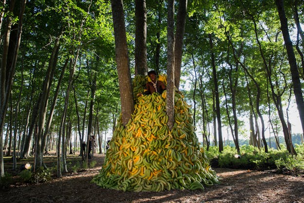 Nile Harris stands towards the top of a pile of yellow and green bananas in a forest of thin trees lit by sunlight. The bananas surround a cluster of four trees at the center of which Nile Harris stands. He is visible from the chest up, each hand clutches a banana, resting on the bananas below him. He is shirtless and wears a noose, the end of which disappears into the bananas. There are production crew members visible in the distant background. 