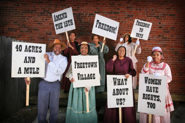 Seven people dressed in old fashioned American clothing stand in front of a brick wall holding signs saying 