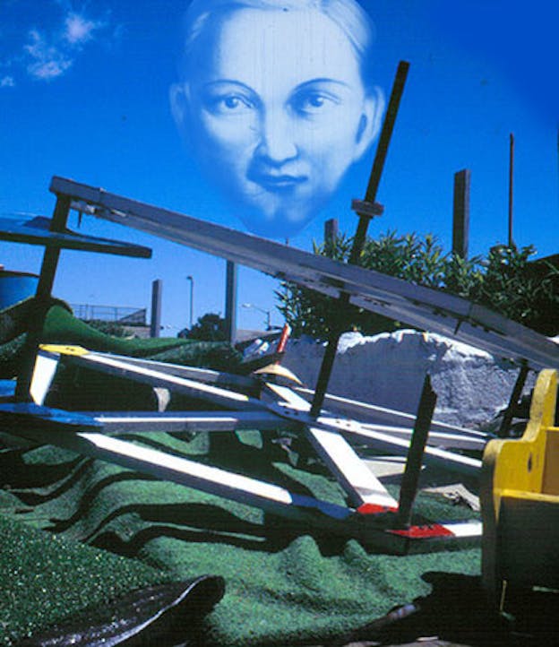 An installation forming a hexagon lies on a grassy green material on the ground.A transparent still film projection of a face is stamped on the top middle of the picture. 