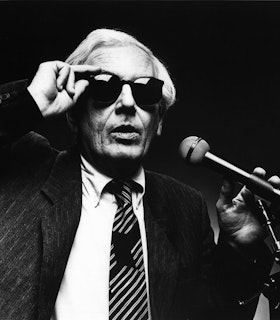A black and white portrait of Robert Ashley wearing a dark suit and tie and black sunglasses. He holds a microphone in his left hand and reaches up to adjust his sunglasses with his right hand. The background is entirely black. 