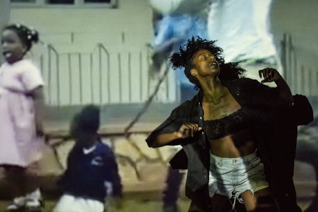 A performer leans backwards, with their arms raised and bent by the shoulders and their eyes closed. They wear a black lace crop top, an unbuttoned black shirt, and jean shorts. A projection of two young girls against a geometric patterned white wall and shorter tan stone wall is over layed across the entire scene. 